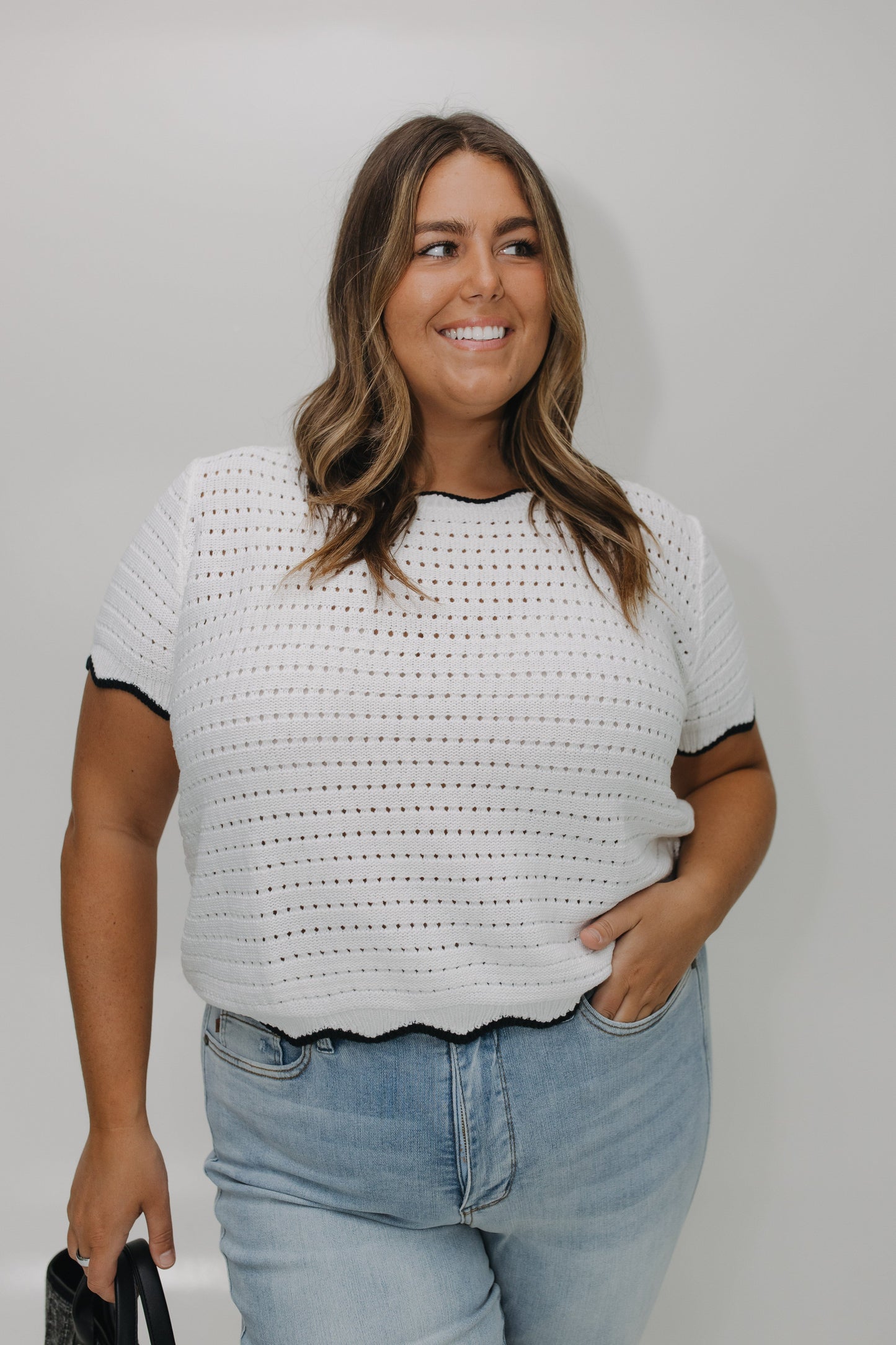 Woman wearing a white crochet top with black trim and blue jeans against a plain background