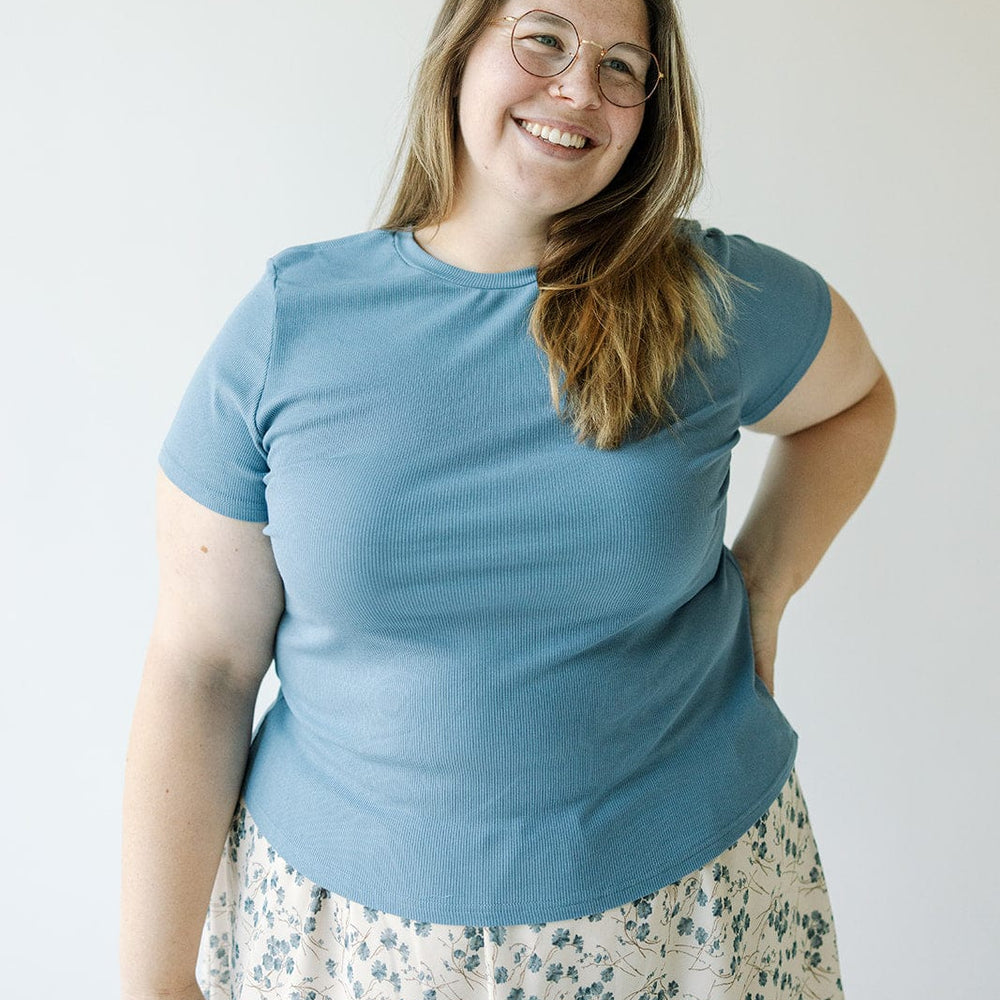 A woman with long hair, wearing glasses, a Mikarose Cropped Short Sleeve Ribbed Tee in Adrift and a floral skirt, stands smiling with one hand on her hip against a plain background.