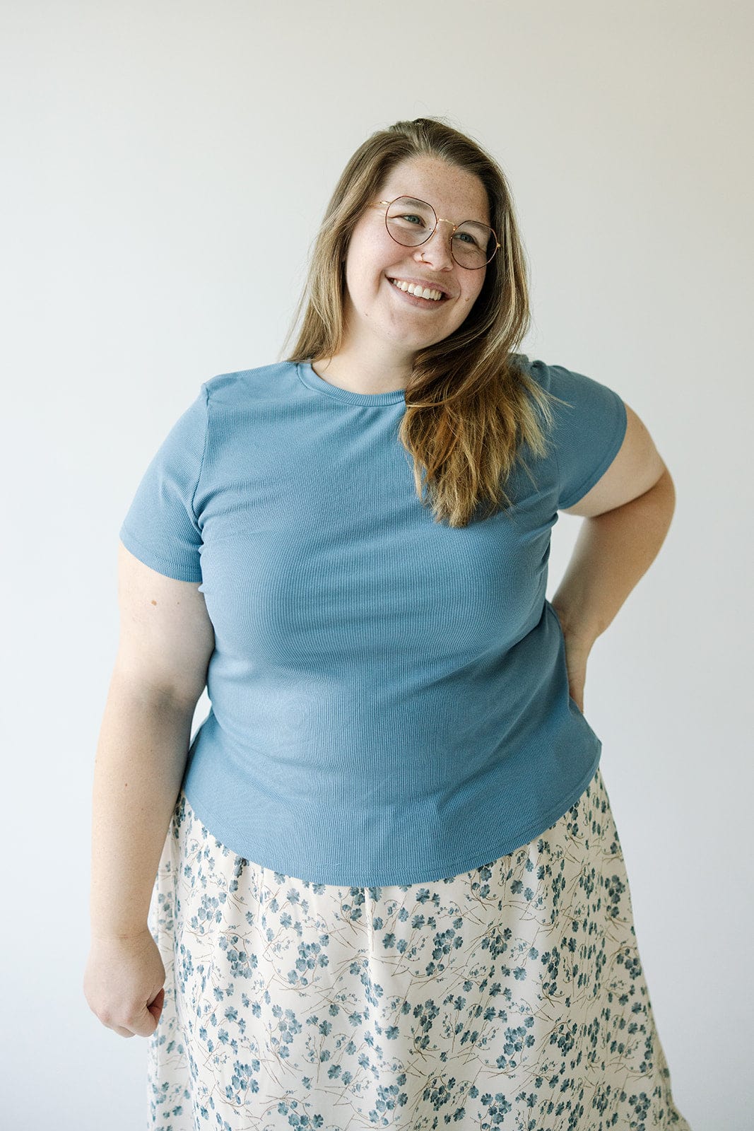 A woman with long hair, wearing glasses, a Mikarose Cropped Short Sleeve Ribbed Tee in Adrift and a floral skirt, stands smiling with one hand on her hip against a plain background.