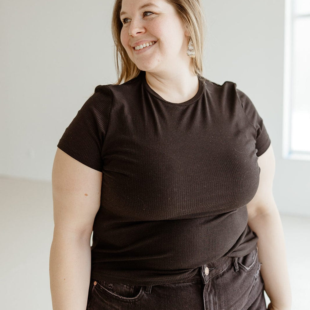 A person with long hair wearing a black ribbed fabric tee and jeans stands indoors, smiling and looking to the side.