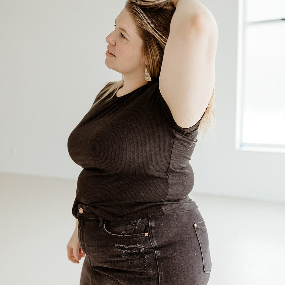 A woman in a black ribbed fabric t-shirt and denim skirt, styled with a long crop length, stands with one hand behind her head in a bright room.