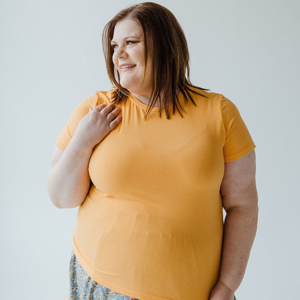 A woman with shoulder-length brown hair smiles while wearing a Love Marlow CROPPED SHORT SLEEVE RIBBED TEE IN CITRUS and a patterned skirt. She is showcasing her unique style against a plain background, the lightweight ribbed fabric adding to her effortless charm.