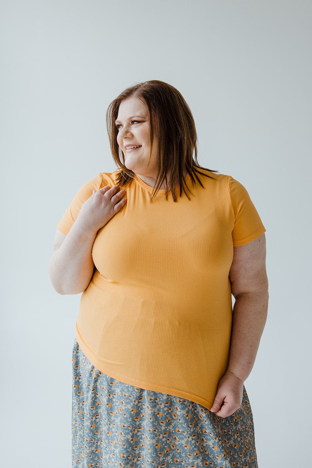 A woman with shoulder-length brown hair smiles while wearing a Love Marlow CROPPED SHORT SLEEVE RIBBED TEE IN CITRUS and a patterned skirt. She is showcasing her unique style against a plain background, the lightweight ribbed fabric adding to her effortless charm.