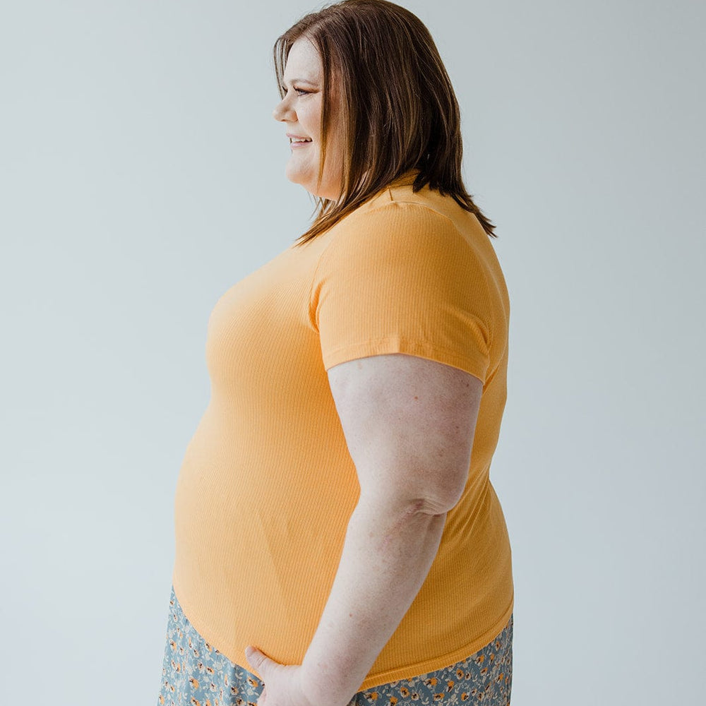 A woman with brown hair stands in profile wearing a CROPPED SHORT SLEEVE RIBBED TEE IN CITRUS by Love Marlow and a patterned skirt, showcasing her unique style against a plain light background.