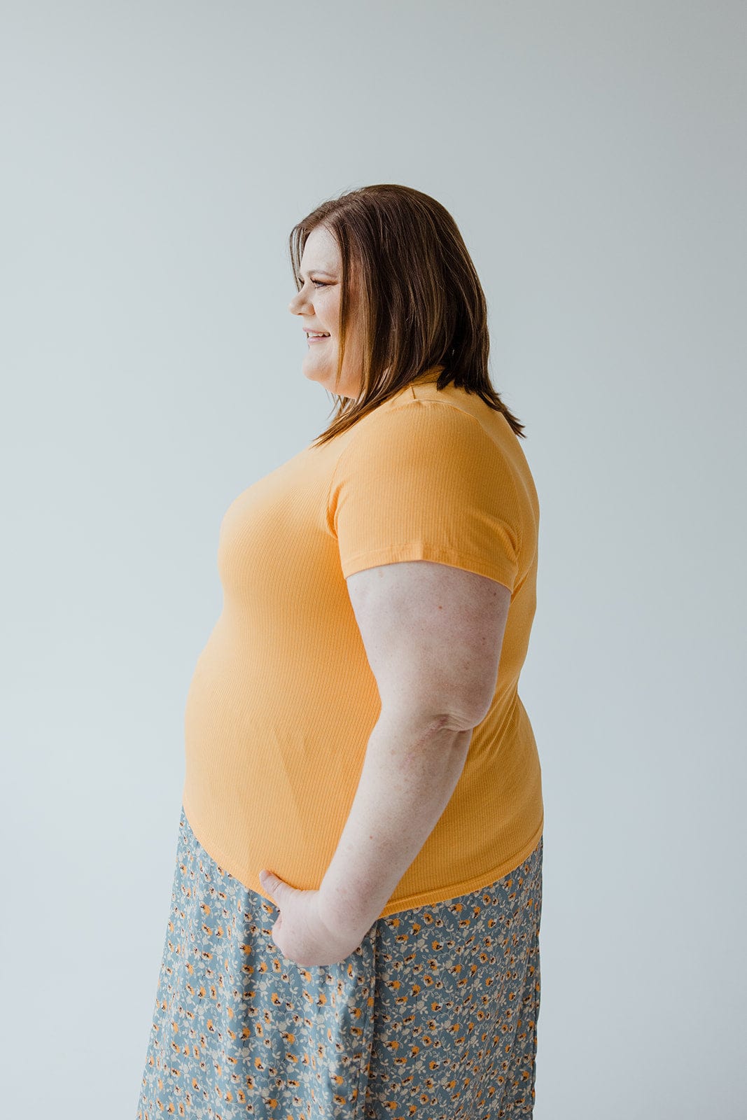 A woman with brown hair stands in profile wearing a CROPPED SHORT SLEEVE RIBBED TEE IN CITRUS by Love Marlow and a patterned skirt, showcasing her unique style against a plain light background.