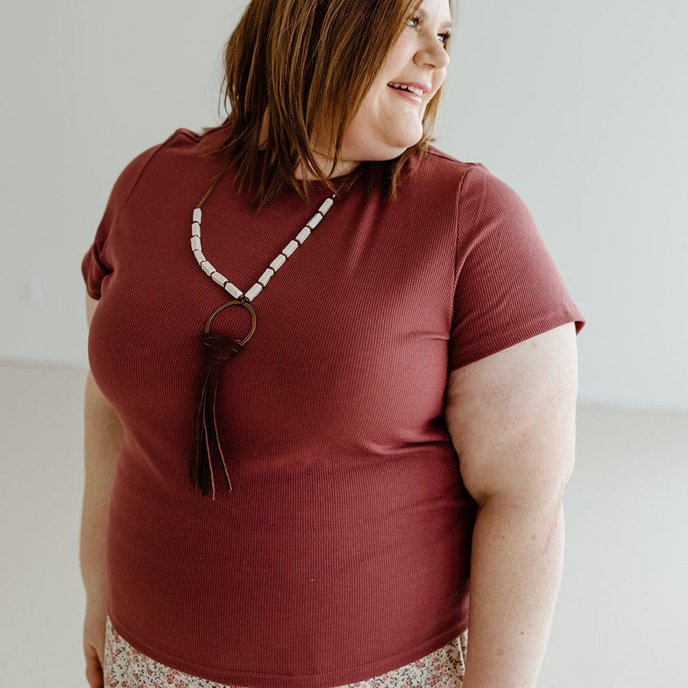 A woman wearing the Mikarose Cropped Short Sleeve Ribbed Tee in Plum and a floral skirt, complemented by a long necklace, glances to the side against a plain background.