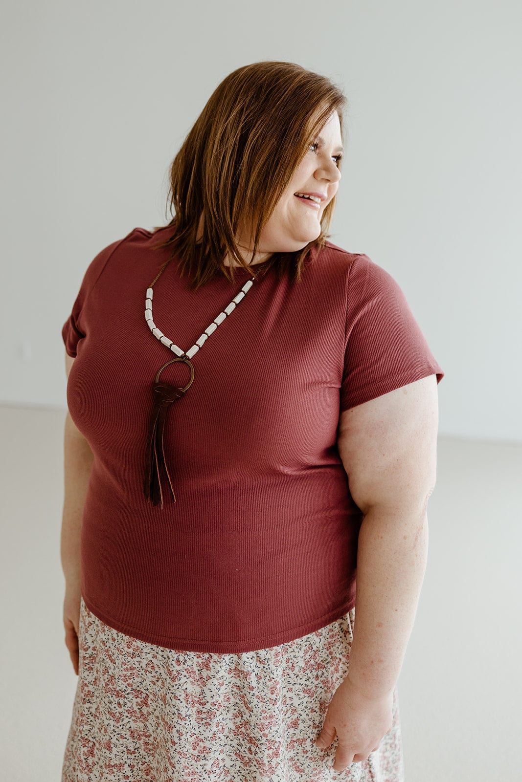 A woman wearing the Mikarose Cropped Short Sleeve Ribbed Tee in Plum and a floral skirt, complemented by a long necklace, glances to the side against a plain background.