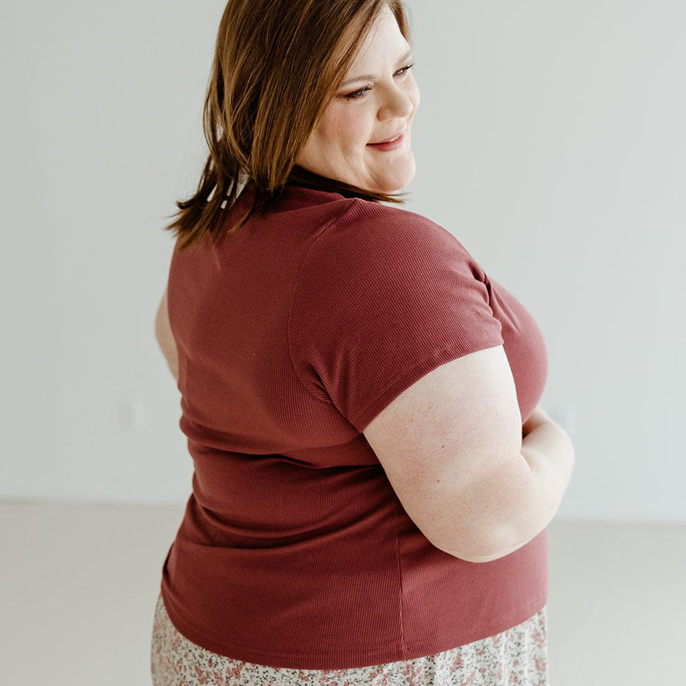 A person wearing Mikarose's Cropped Short Sleeve Ribbed Tee in plum and a floral skirt stands smiling, looking over their shoulder against a plain background.