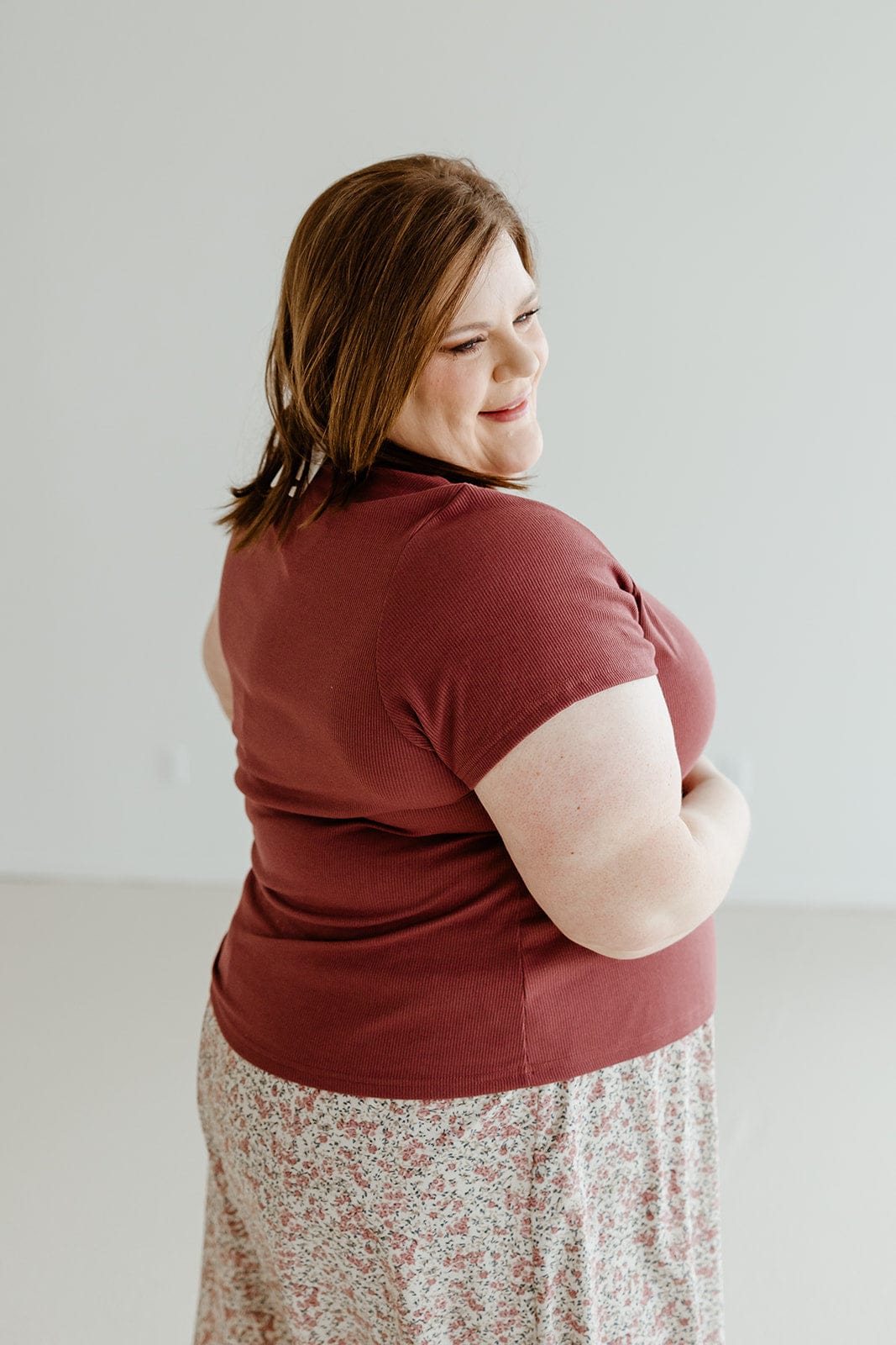 A person wearing Mikarose's Cropped Short Sleeve Ribbed Tee in plum and a floral skirt stands smiling, looking over their shoulder against a plain background.