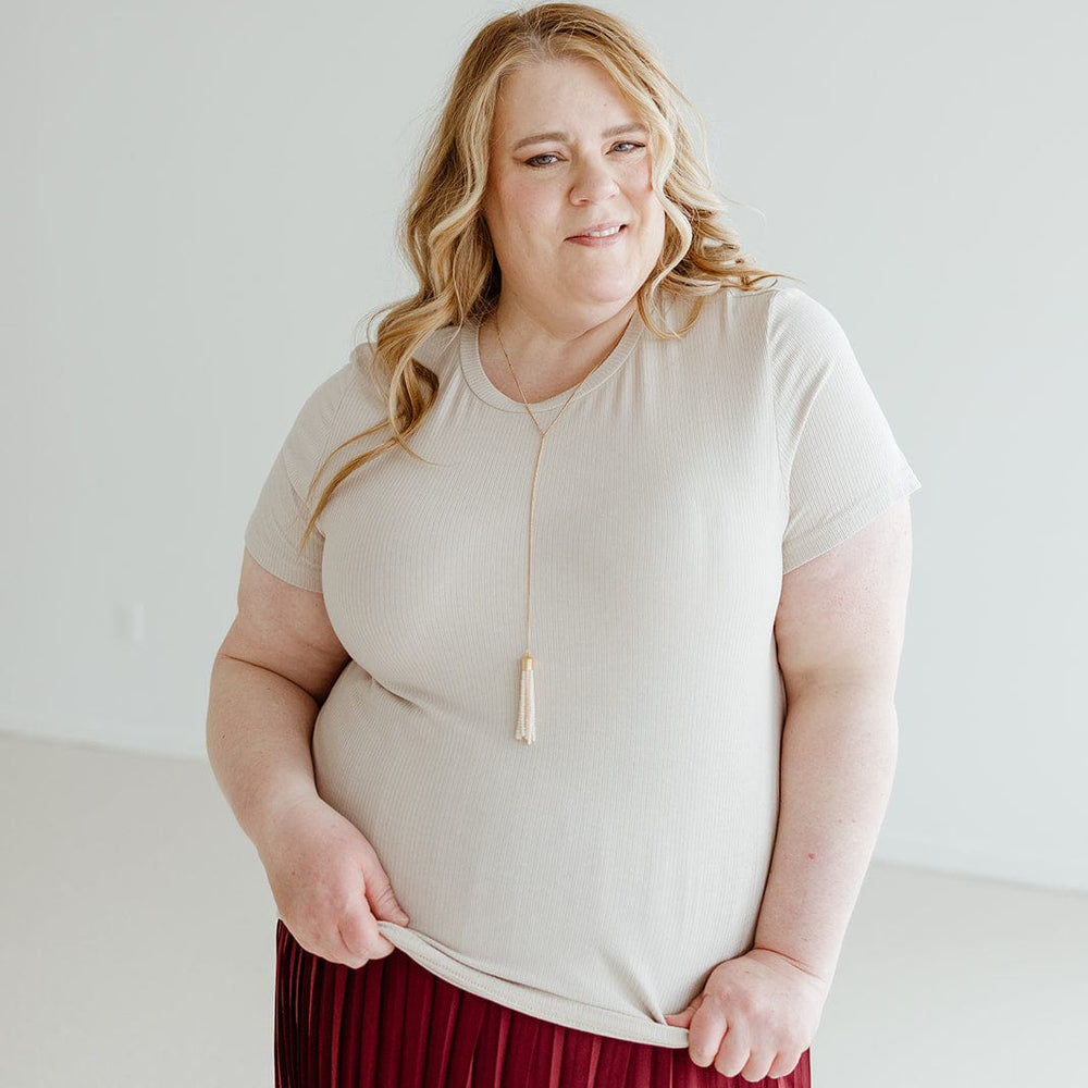 A person with long, wavy hair wearing a versatile neutral CROPPED SHORT SLEEVE RIBBED TEE IN SAND by Love Marlow, a red pleated skirt, and a necklace with a tassel stands against a plain light background.