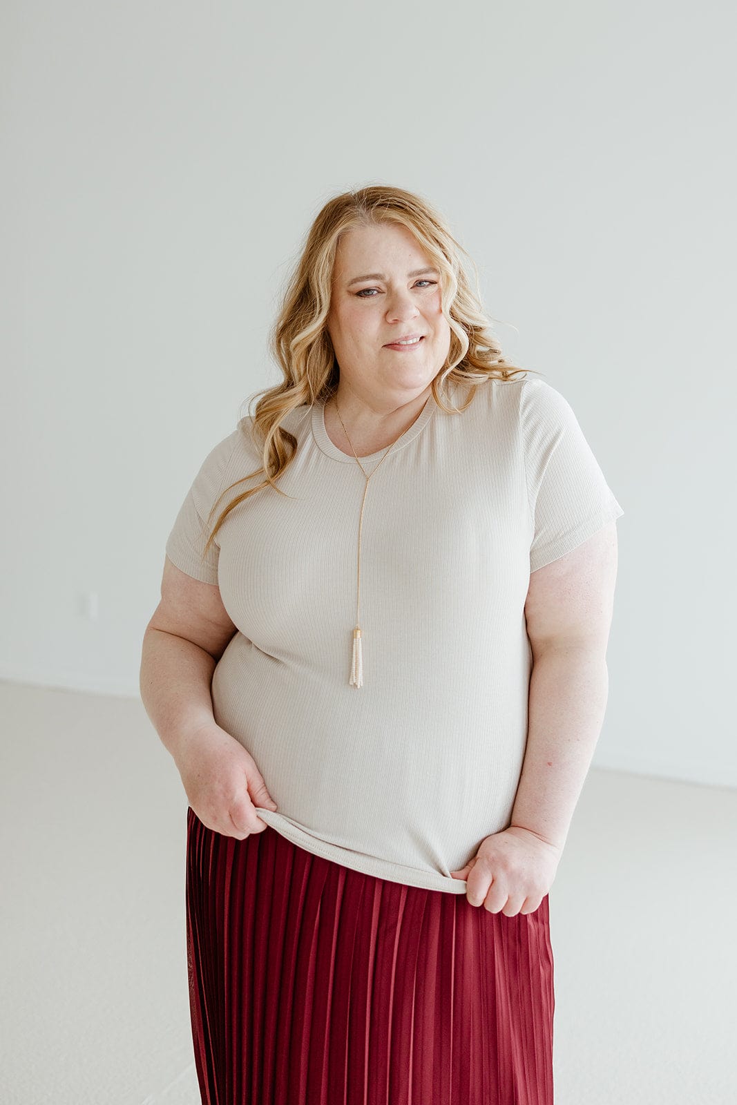 A person with long, wavy hair wearing a versatile neutral CROPPED SHORT SLEEVE RIBBED TEE IN SAND by Love Marlow, a red pleated skirt, and a necklace with a tassel stands against a plain light background.