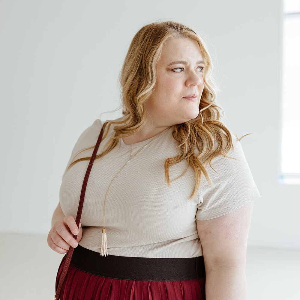 A woman with long blonde hair, wearing a Love Marlow CROPPED SHORT SLEEVE RIBBED TEE IN SAND and a red pleated skirt, stands indoors looking to the side. She has a crossbody bag, and there's a window in the background. Her lightweight textured fabric outfit adds to her effortless style.