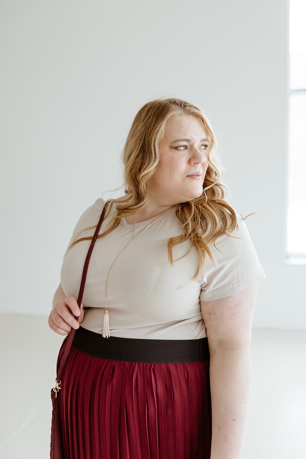 A woman with long blonde hair, wearing a Love Marlow CROPPED SHORT SLEEVE RIBBED TEE IN SAND and a red pleated skirt, stands indoors looking to the side. She has a crossbody bag, and there's a window in the background. Her lightweight textured fabric outfit adds to her effortless style.