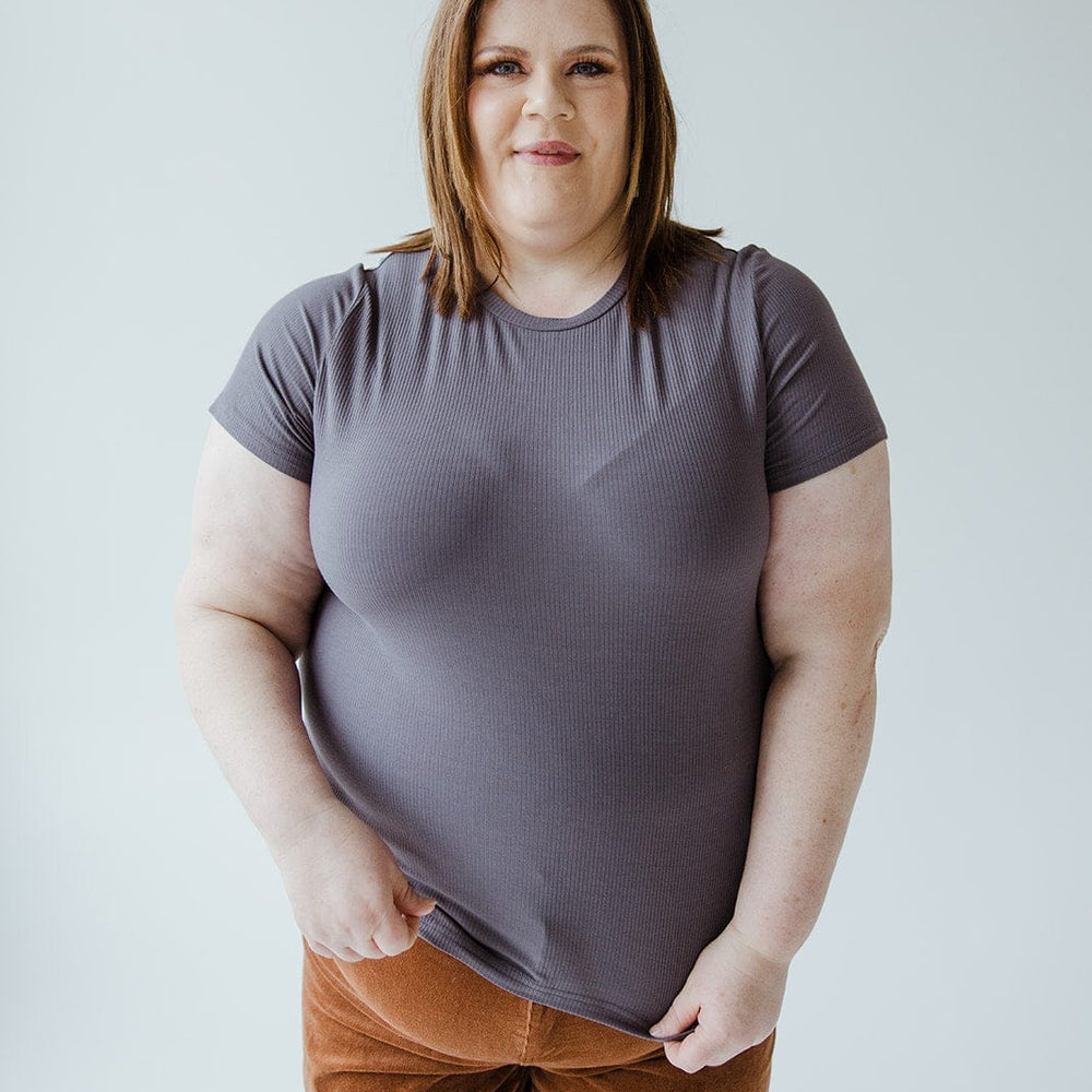 A person with shoulder-length brown hair wearing the Love Marlow CROPPED SHORT SLEEVE RIBBED TEE IN STORMY and brown pants stands against a plain background.