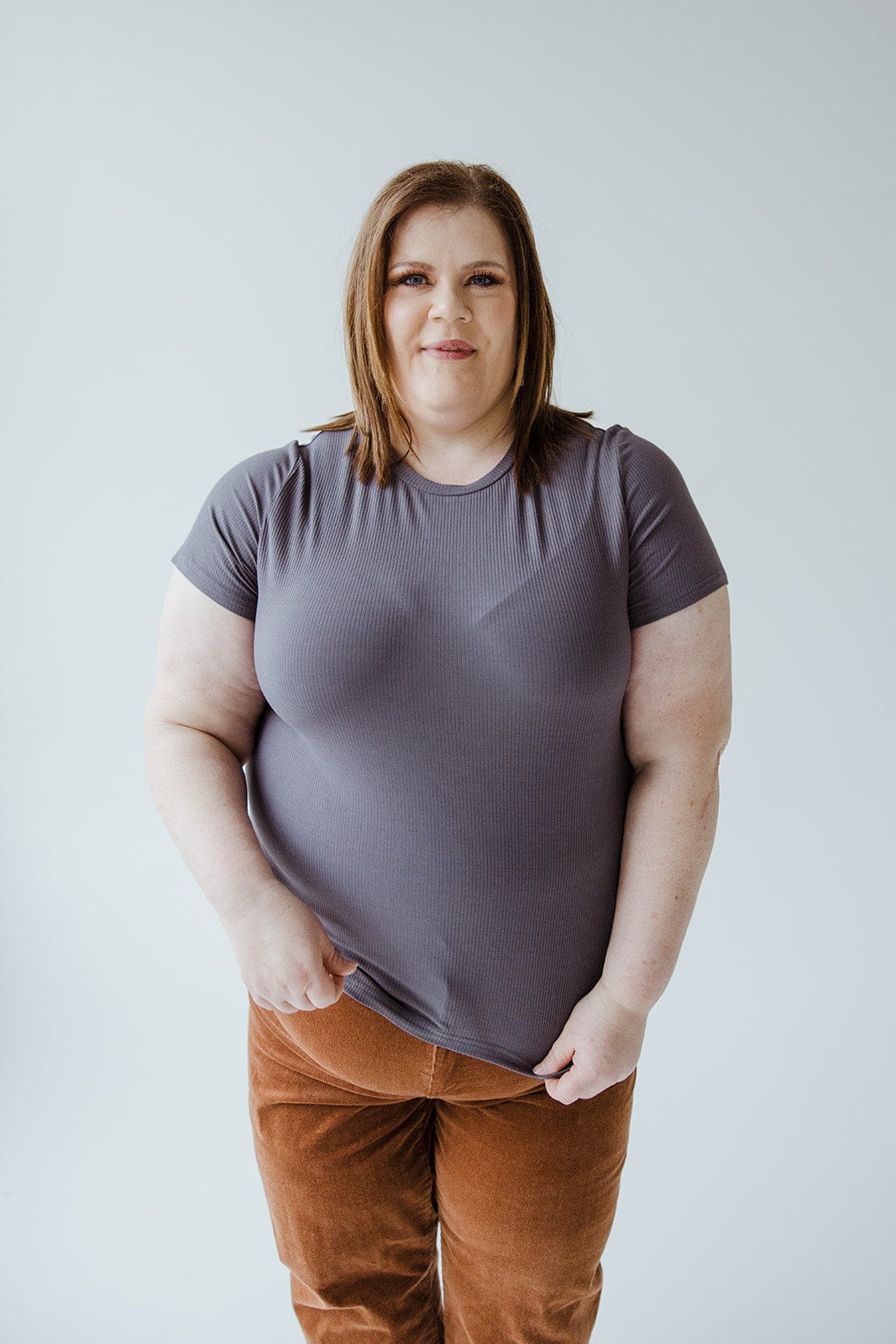 A person with shoulder-length brown hair wearing the Love Marlow CROPPED SHORT SLEEVE RIBBED TEE IN STORMY and brown pants stands against a plain background.