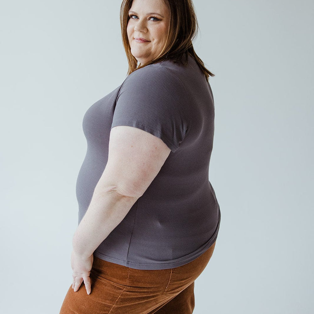 A person with brown hair, wearing the CROPPED SHORT SLEEVE RIBBED TEE IN STORMY from Love Marlow and brown pants, stands and looks over their shoulder against a plain light background.