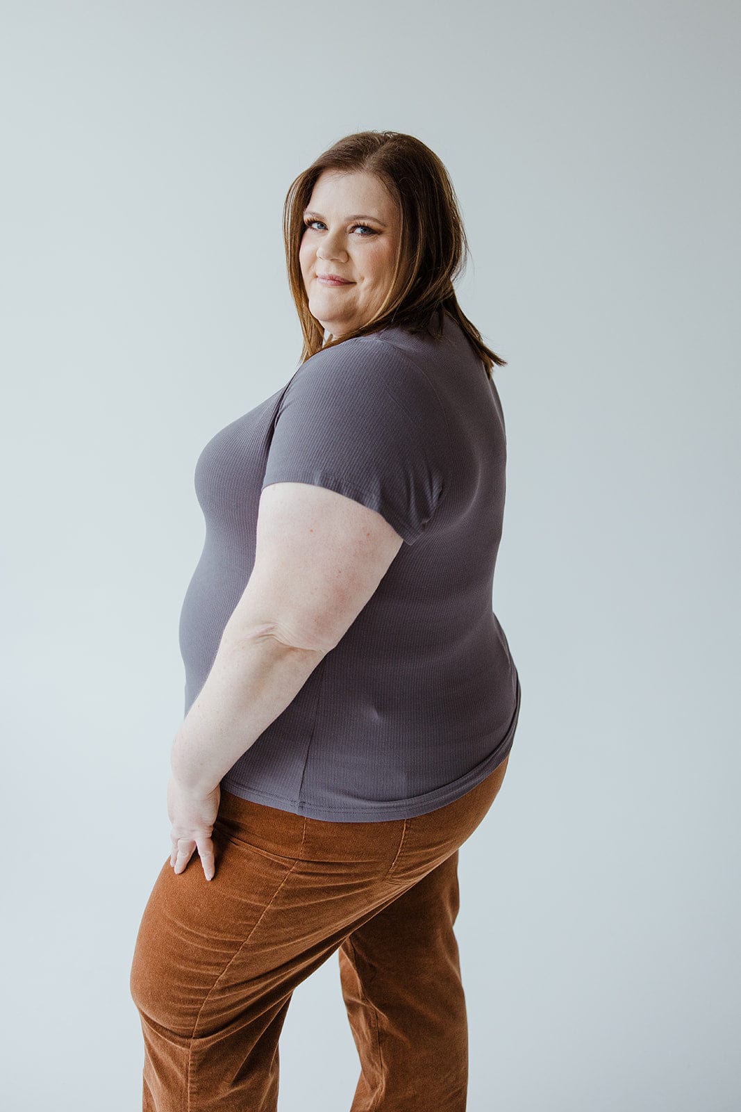 A person with brown hair, wearing the CROPPED SHORT SLEEVE RIBBED TEE IN STORMY from Love Marlow and brown pants, stands and looks over their shoulder against a plain light background.