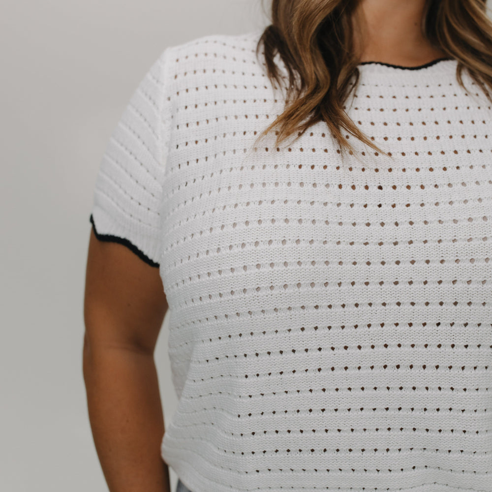 Woman wearing a white crochet top with black trim against a plain background