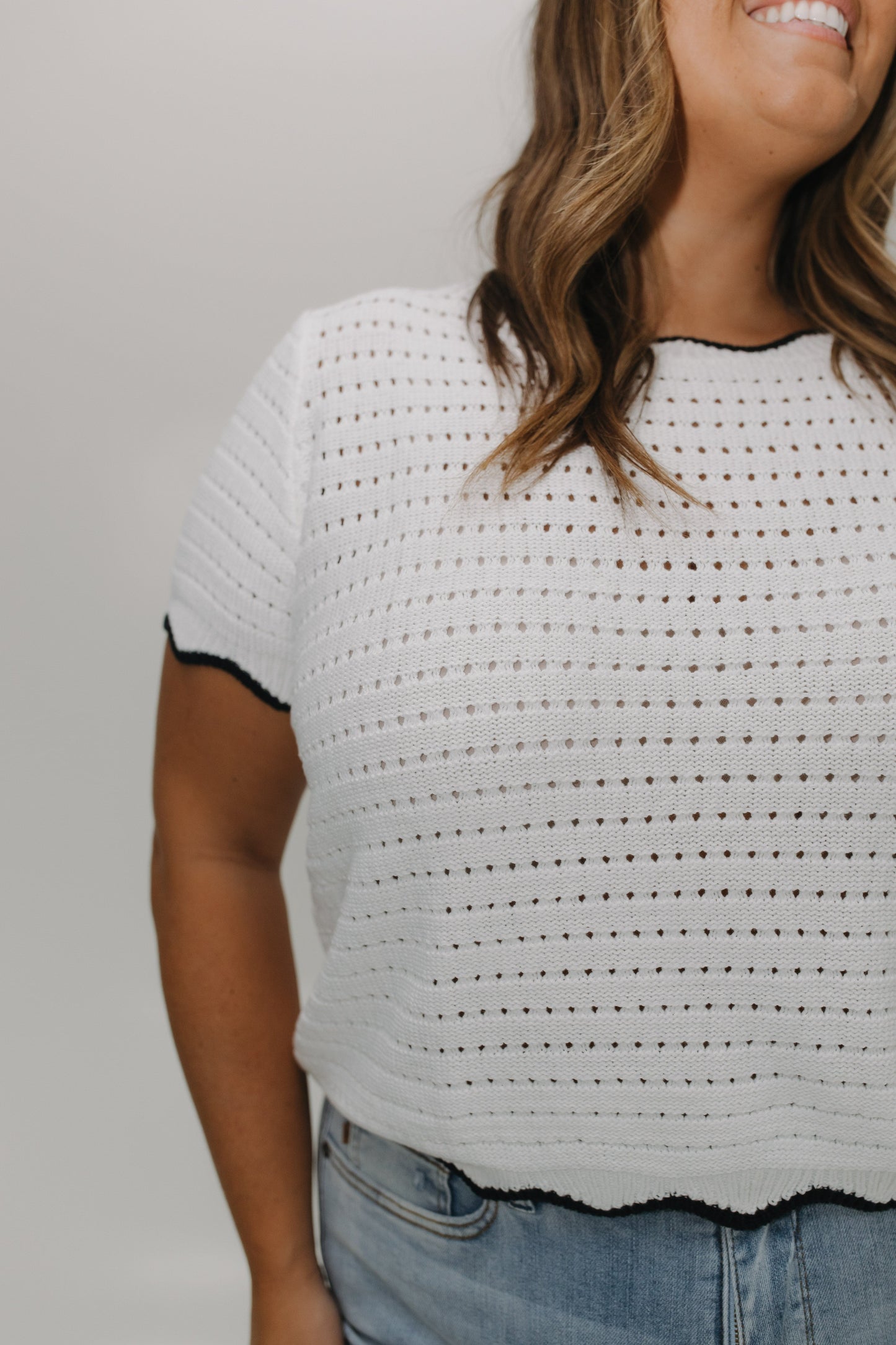 Woman wearing a white crochet top with black trim against a plain background