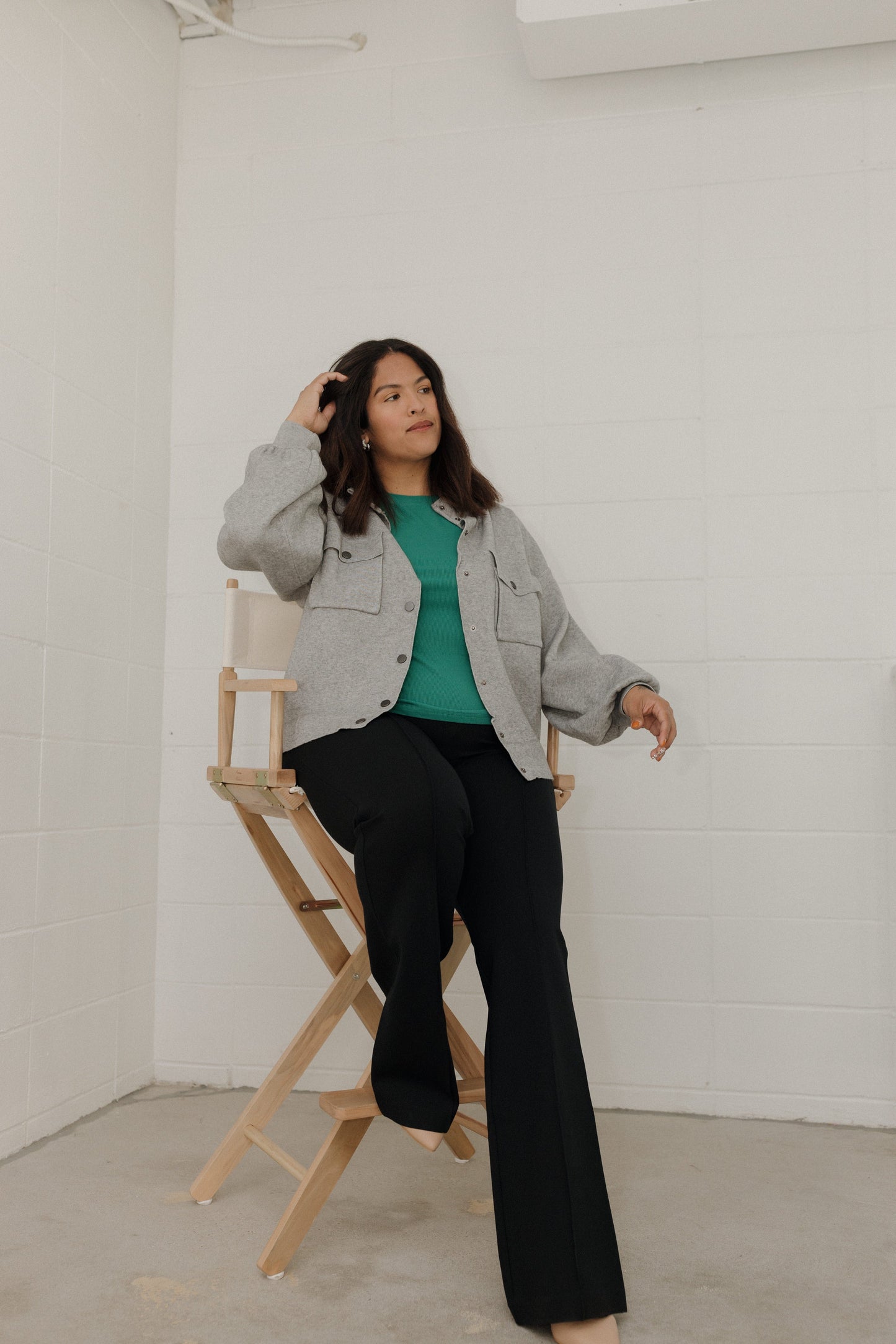 Woman sitting on a director's chair wearing a gray jacket, green shirt, and black pants in a minimalistic setting.
