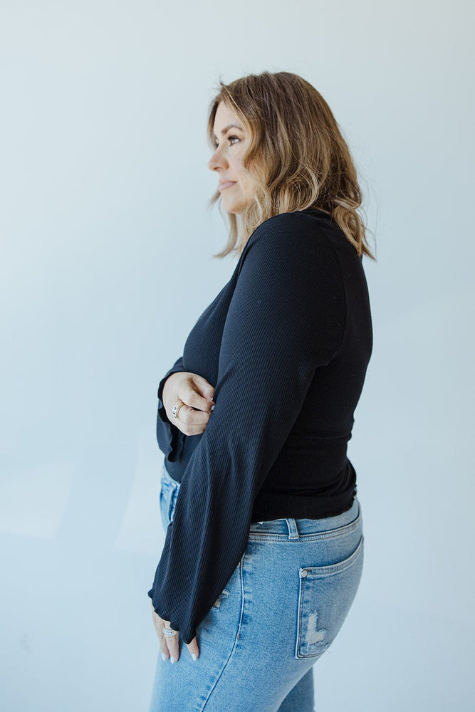 Wearing a Mikarose Cropped Long Sleeve Ribbed Tee in black and blue jeans, the person stands against a light background, gazing to the side.