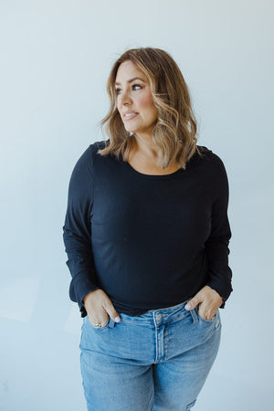 A person dressed in a black, ribbed Mikarose cropped long sleeve tee and blue jeans stands against a light background, looking to the right with hands in pockets.