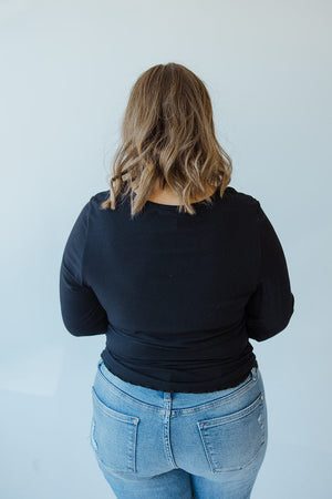 A person with shoulder-length wavy hair, wearing the Mikarose Cropped Long Sleeve Ribbed Tee in black and blue jeans, stands with their back to the camera.