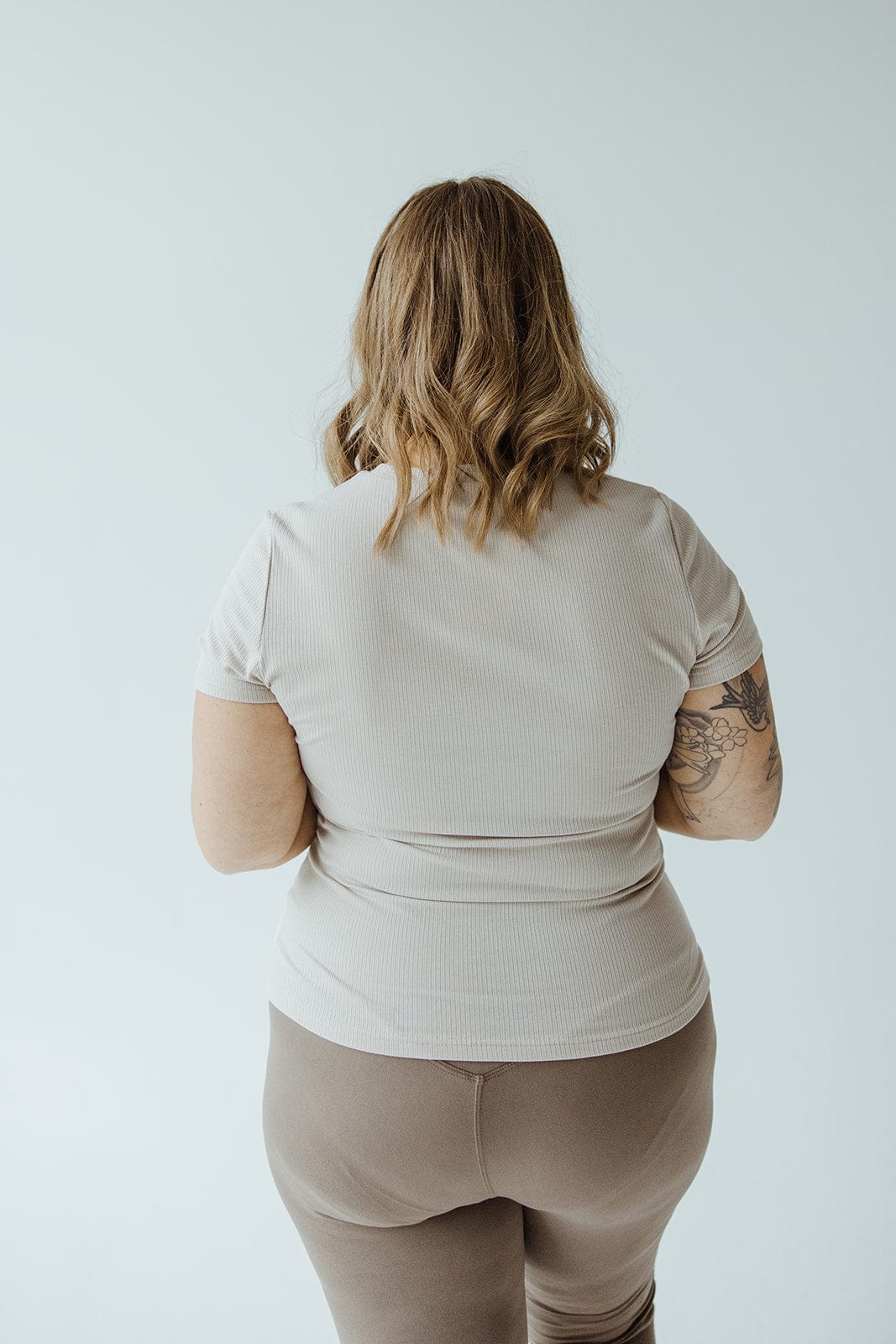 A person with shoulder-length hair, wearing a light-colored short-sleeve shirt and pants, stands with their back to the camera. The shirt offers a lightweight feel perfect for layering options, while a floral tattoo adorns their arm.