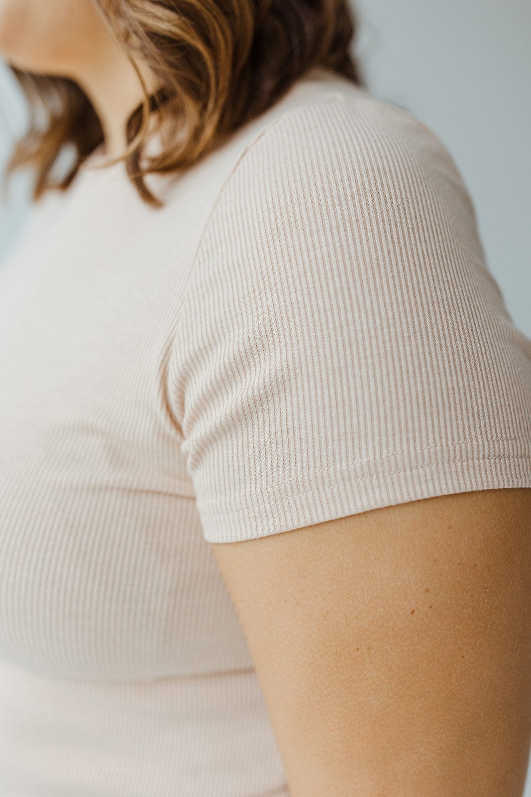 Close-up of a person wearing an oatmeal cropped tee with ribbed short sleeves, complemented by their shoulder-length brown hair.