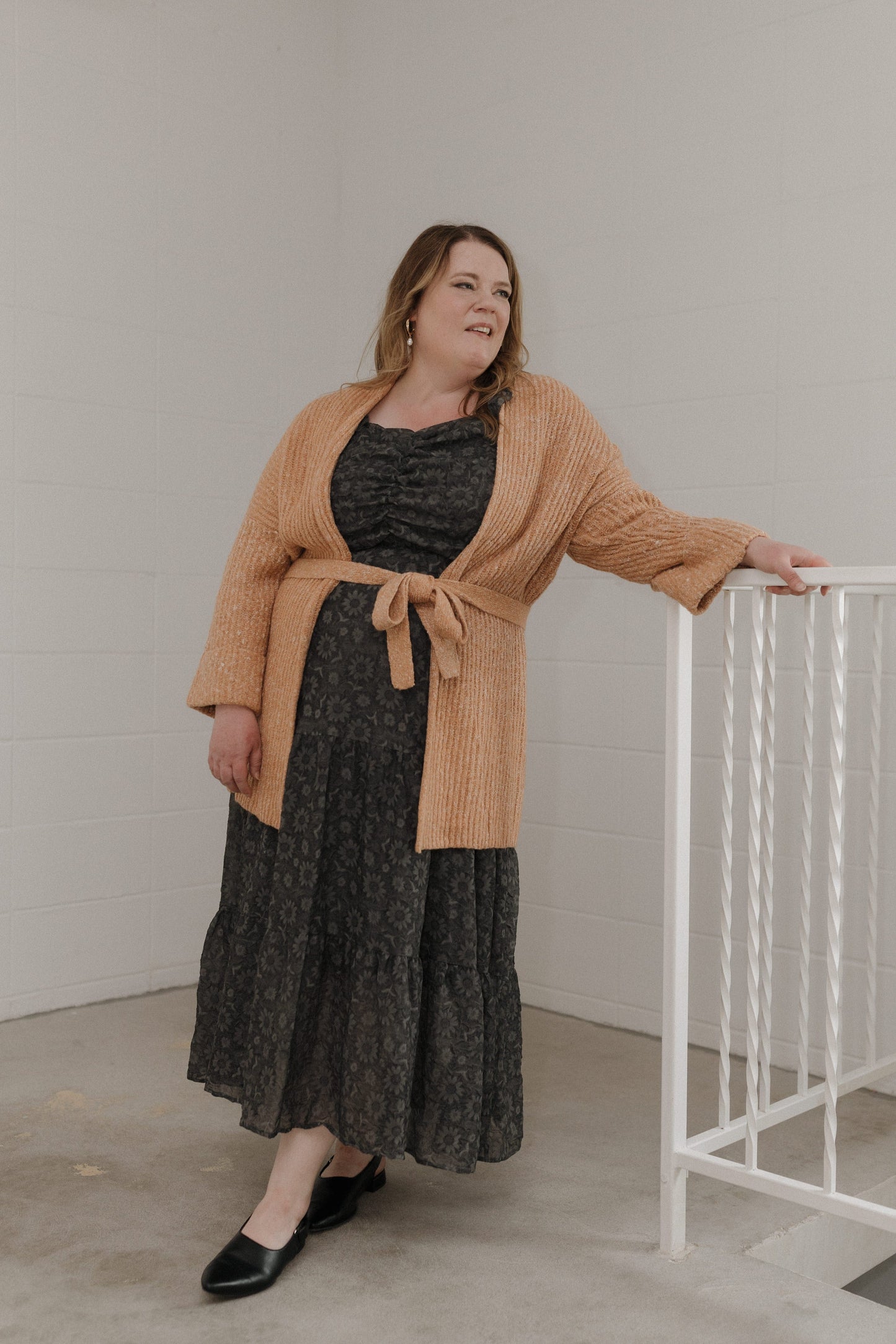 Woman wearing a brown cardigan over a black dress, standing indoors against a plain wall.