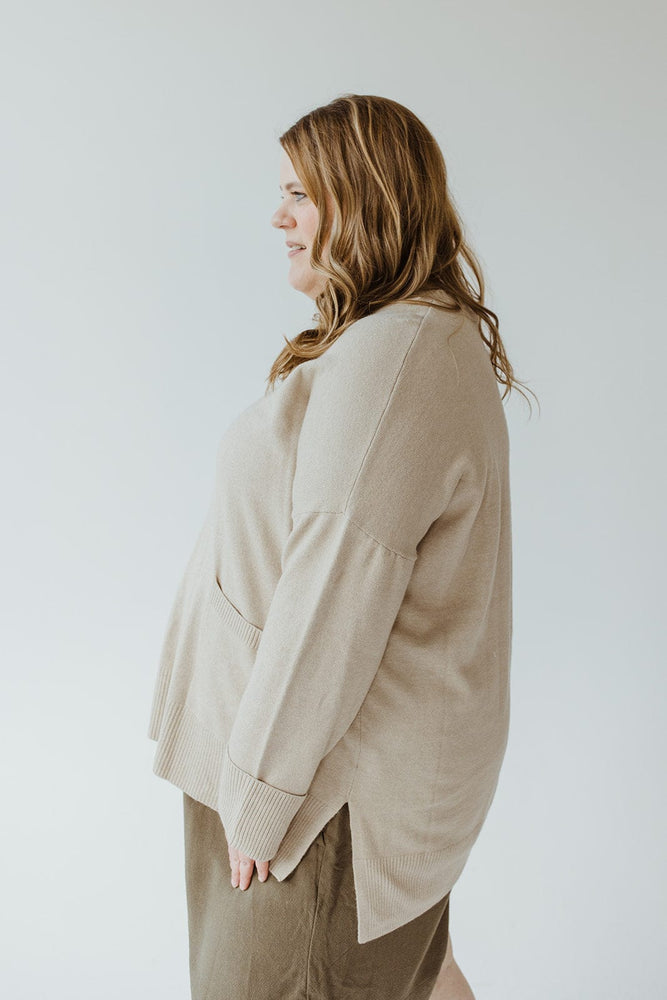 A woman with long hair is wearing a classic piece from Yesta, the CUFFED SWEATER WITH POCKETS in beige, paired with an olive skirt, standing in profile against a plain background.