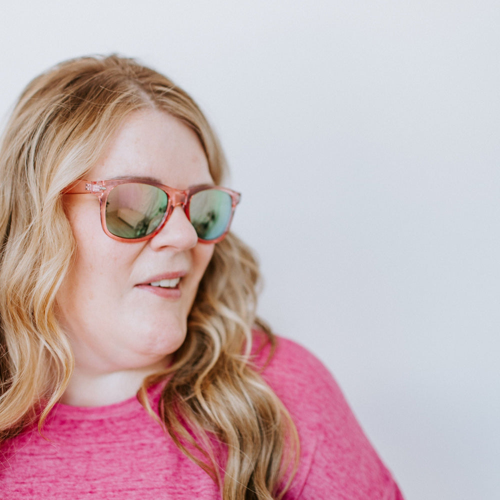 A woman with long blonde hair, wearing a pink shirt and Love Marlow CURACAO SUNGLASSES IN FLAMINGO with UVA/UVB protection, looks to the side against a plain background.