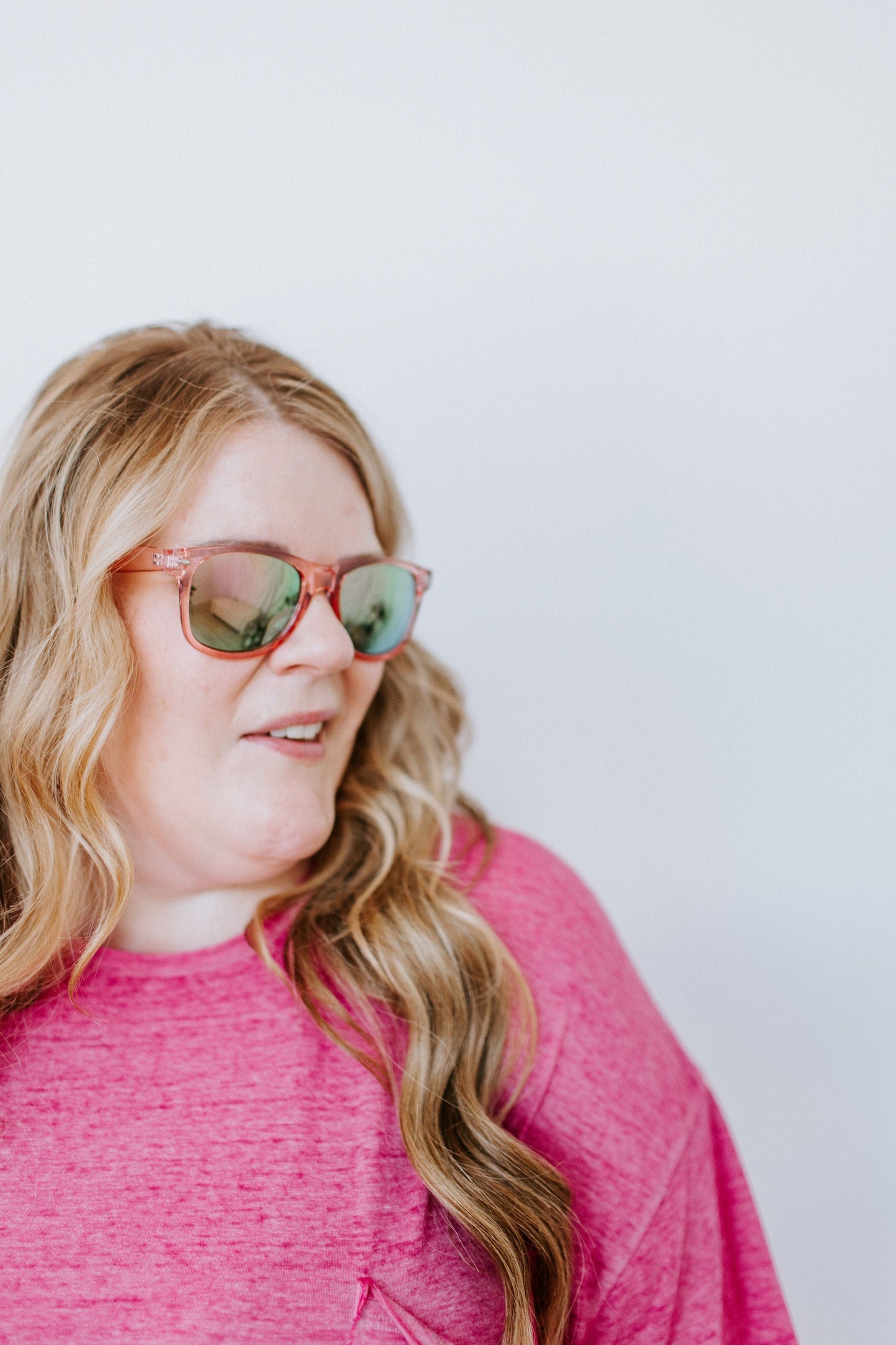 A woman with long blonde hair, wearing a pink shirt and Love Marlow CURACAO SUNGLASSES IN FLAMINGO with UVA/UVB protection, looks to the side against a plain background.