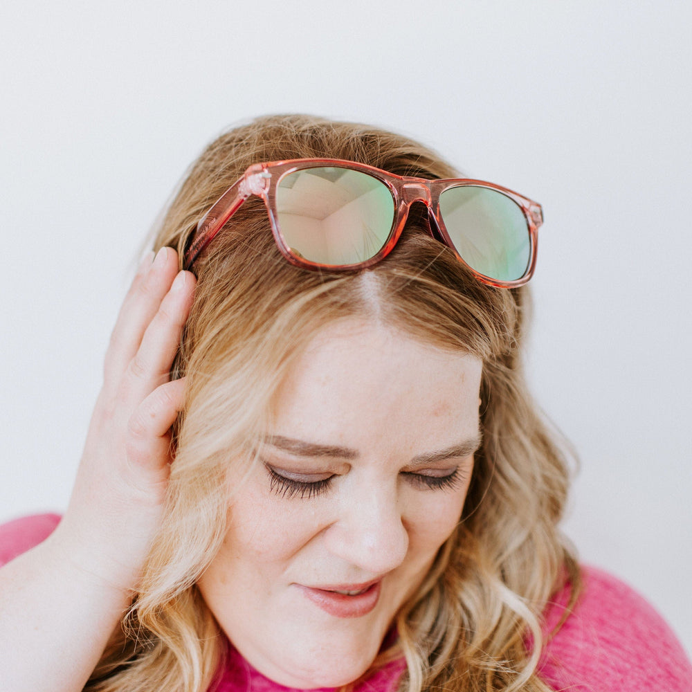 Person with wavy hair wearing a pink top, tilting their head and touching their face, smiling with eyes closed. CURACAO SUNGLASSES IN FLAMINGO by Love Marlow rest stylishly on their head, offering UVA/UVB protection.