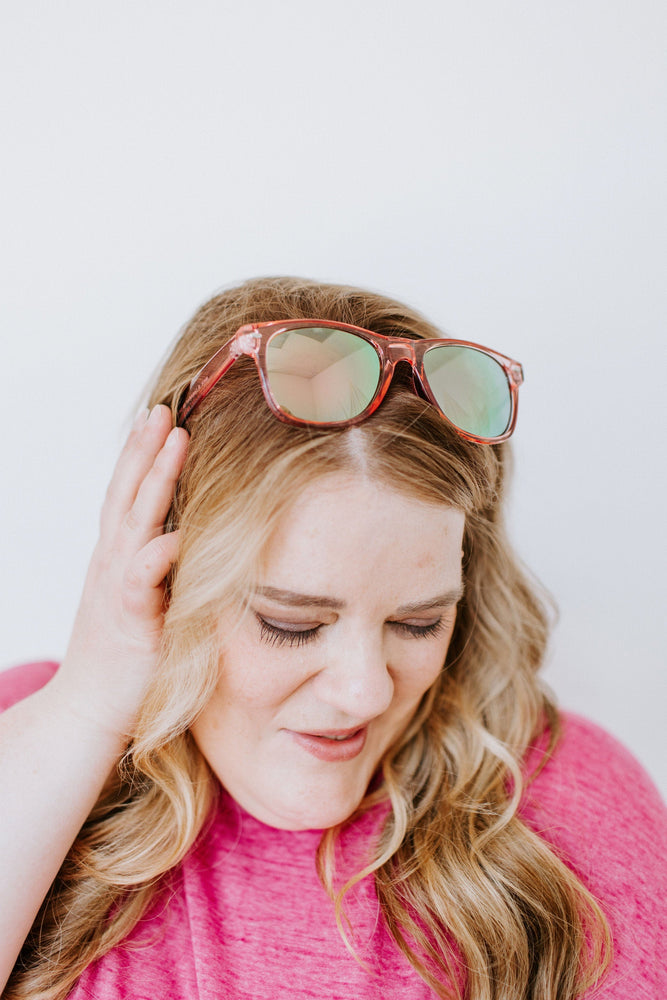 Person with wavy hair wearing a pink top, tilting their head and touching their face, smiling with eyes closed. CURACAO SUNGLASSES IN FLAMINGO by Love Marlow rest stylishly on their head, offering UVA/UVB protection.