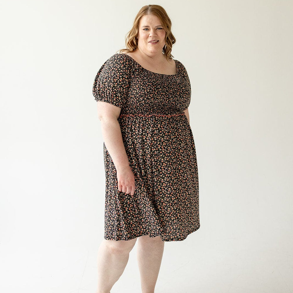 Woman in a DAINTY FLORAL SMOCKED BODICE DRESS by Love Marlow stands against a plain, white background, looking at the camera with a neutral expression. She is wearing light-colored sandals.