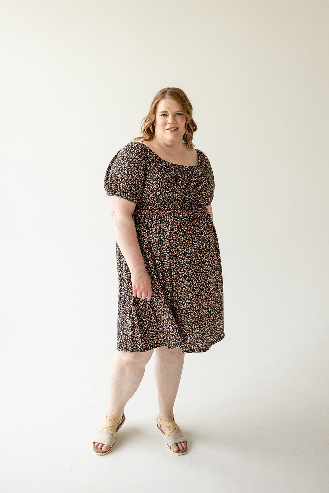 Woman in a DAINTY FLORAL SMOCKED BODICE DRESS by Love Marlow stands against a plain, white background, looking at the camera with a neutral expression. She is wearing light-colored sandals.