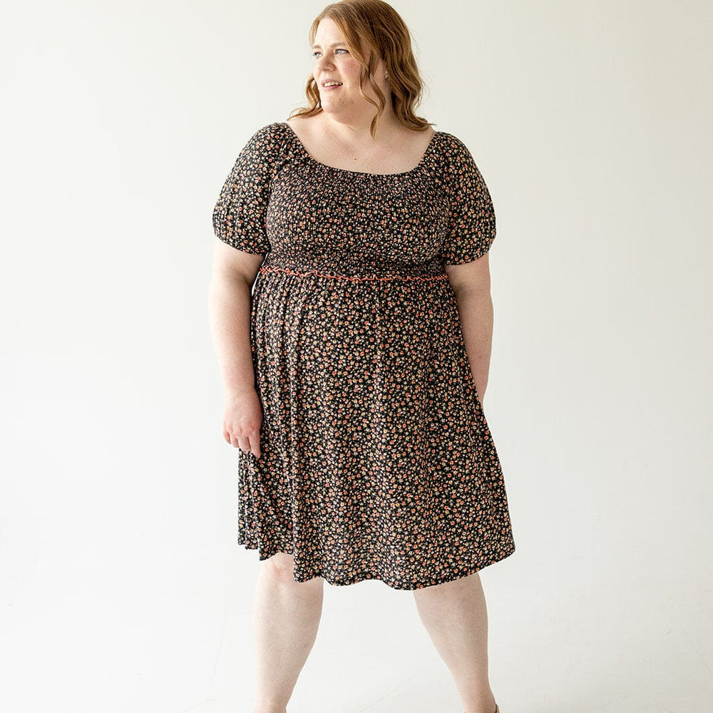 A woman in a Love Marlow DAINTY FLORAL SMOCKED BODICE DRESS with an A-line skirt and beige sandals stands against a plain white background, looking to her left with a smile.