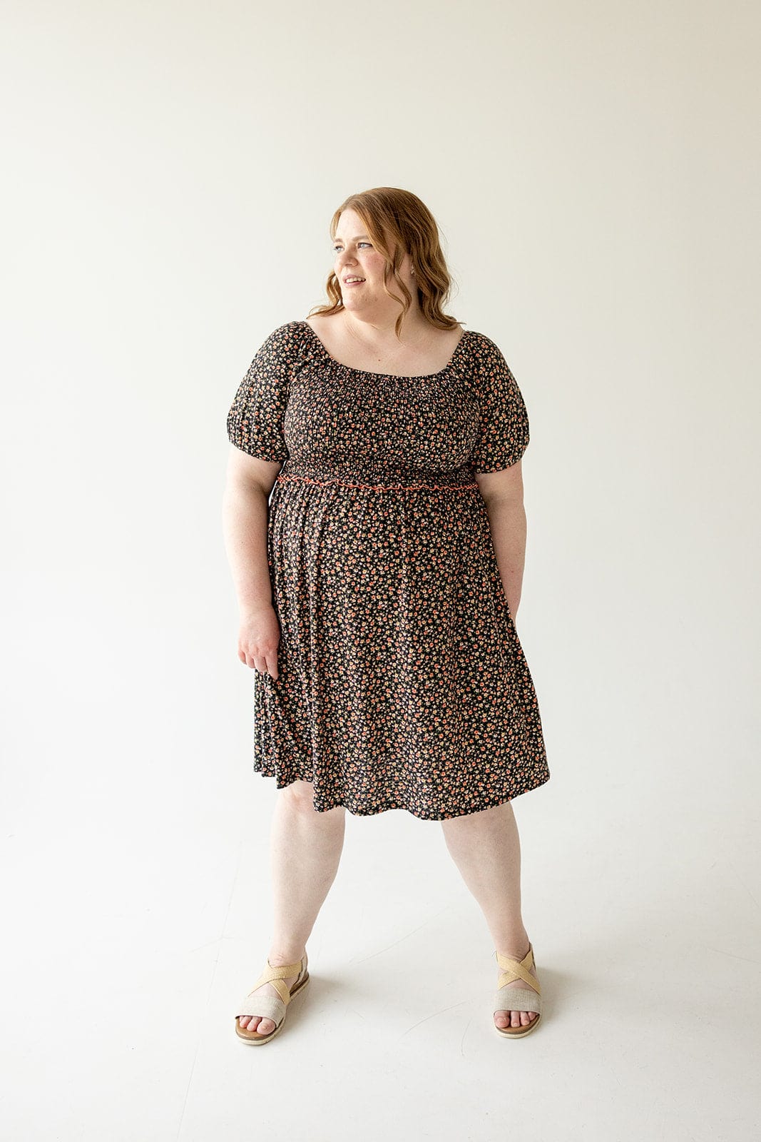 A woman in a Love Marlow DAINTY FLORAL SMOCKED BODICE DRESS with an A-line skirt and beige sandals stands against a plain white background, looking to her left with a smile.