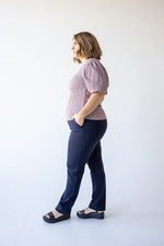 A woman with light brown hair, wearing a purple textured blouse and luxurious fabric dress pants, stands facing away from the camera against a plain background.