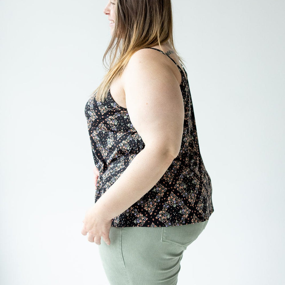 A woman stands sideways, wearing a black diamond-patterned floral cami with a V-neckline and light green pants against a plain background.