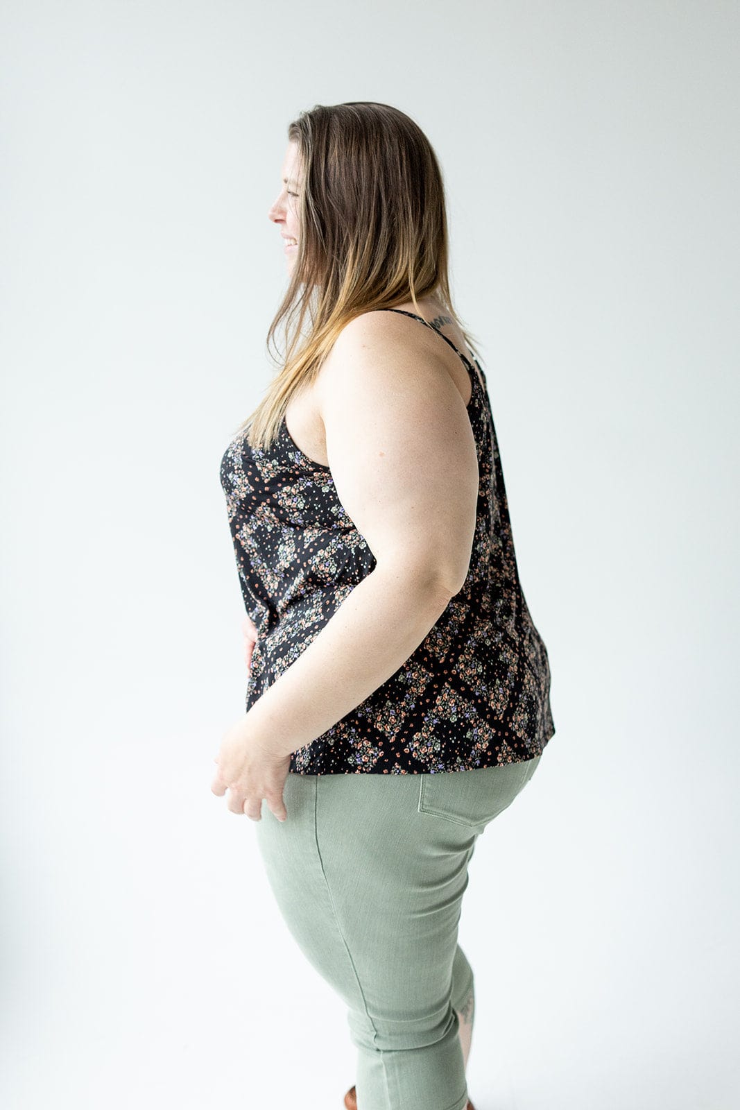 A woman stands sideways, wearing a black diamond-patterned floral cami with a V-neckline and light green pants against a plain background.