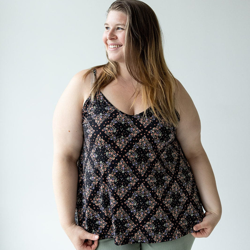 A person stands against a plain backdrop, smiling and wearing a diamond patterned floral cami with adjustable straps, paired with green pants.