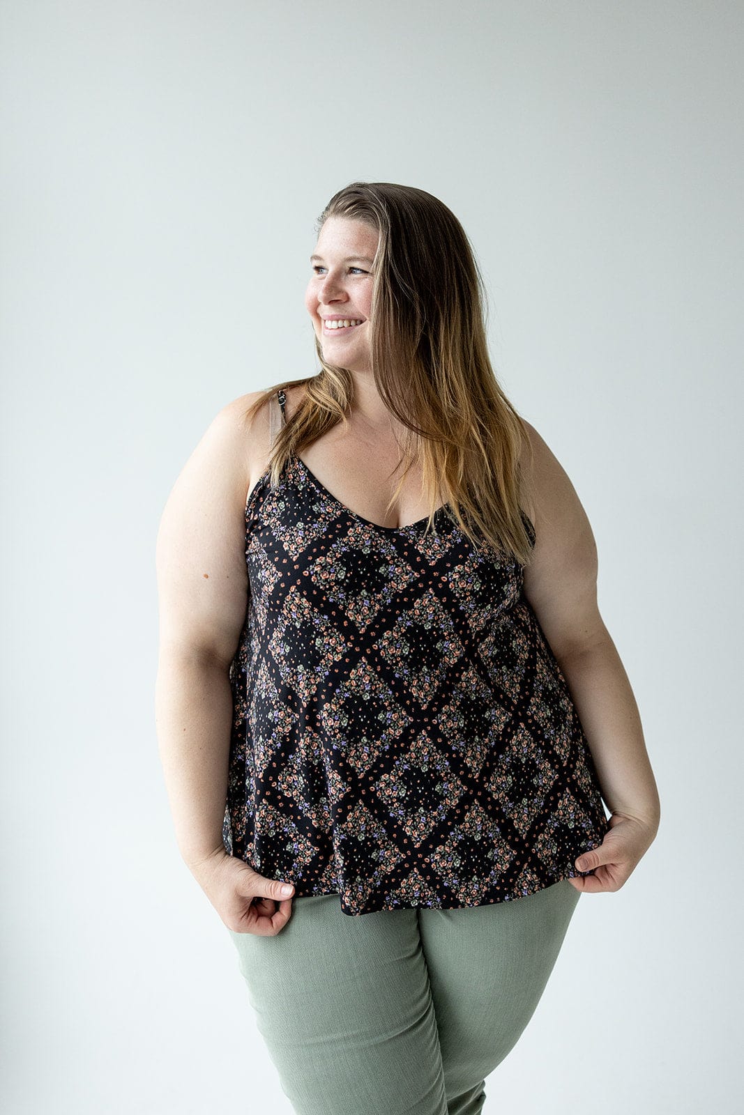 A person stands against a plain backdrop, smiling and wearing a diamond patterned floral cami with adjustable straps, paired with green pants.