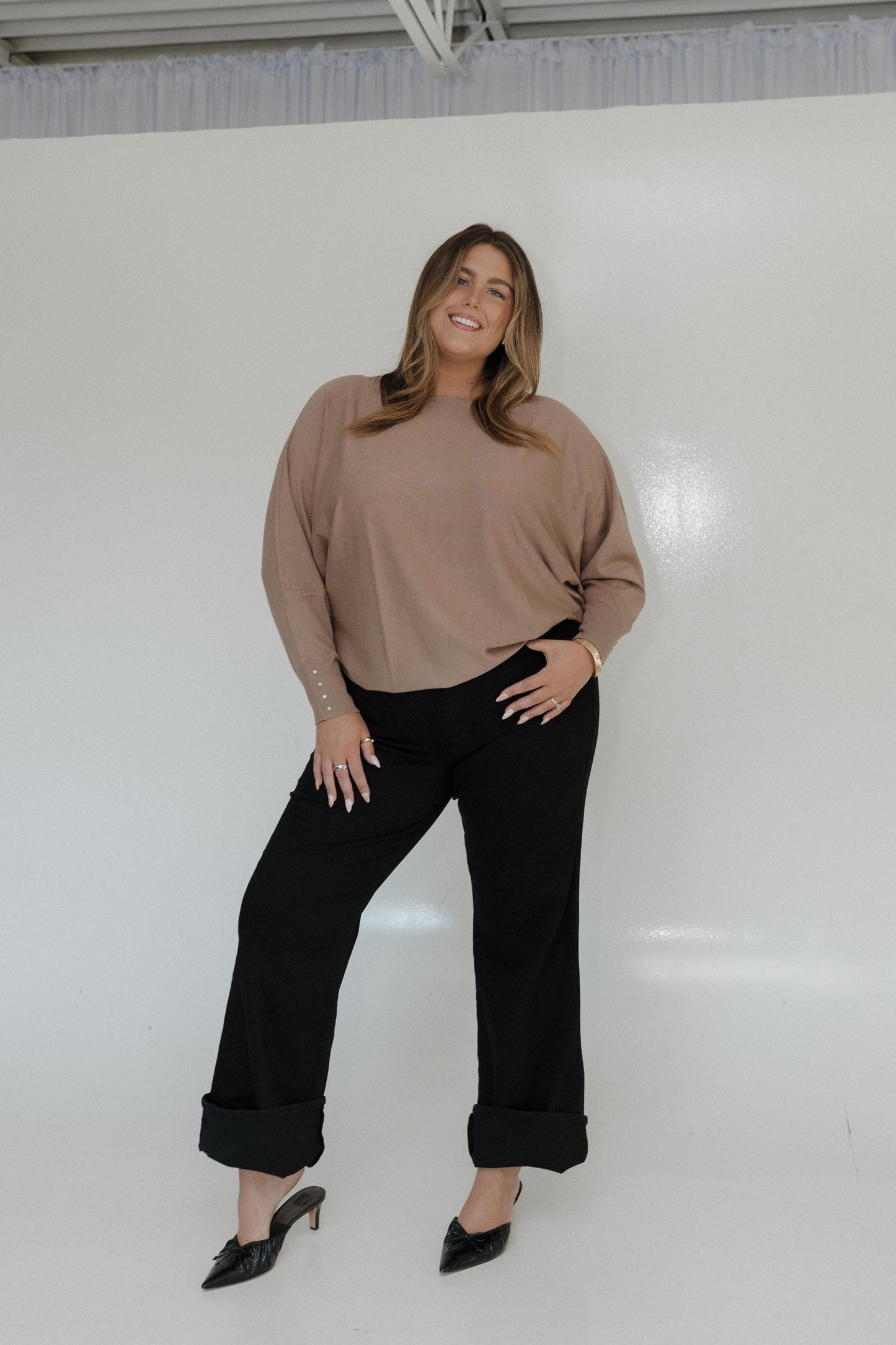 Woman wearing a beige sweater and black pants against a white wall