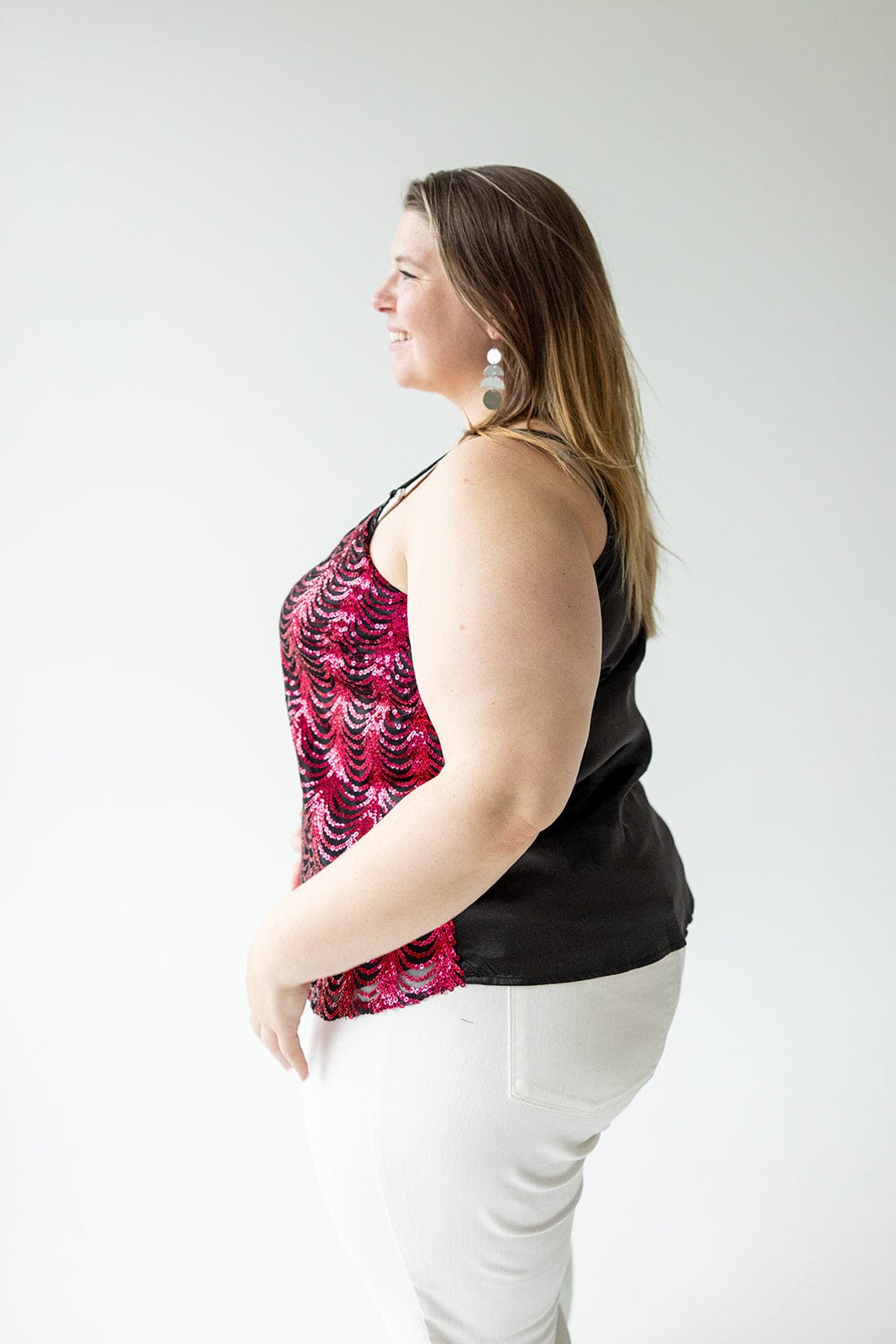 A person with long hair, wearing a sleeveless red and black top featuring adjustable straps and white pants, stands in profile against a plain white background.