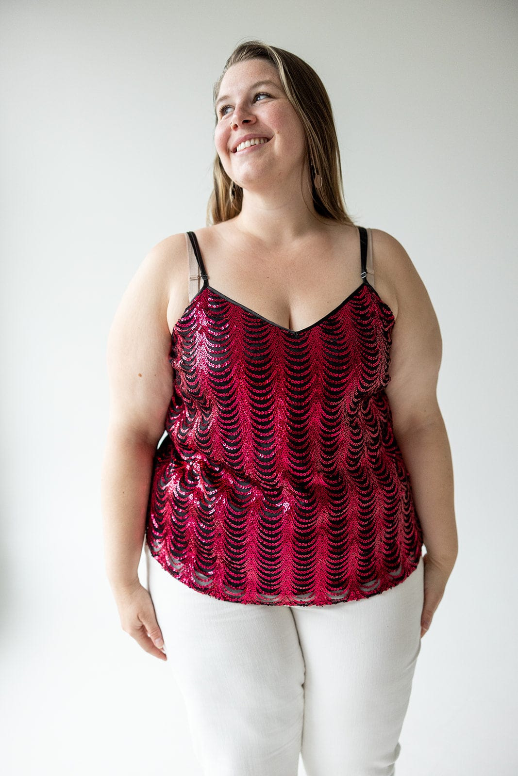 A woman in a hot pink, double V-neck sequin cami with adjustable straps and white pants smiles against a plain background.