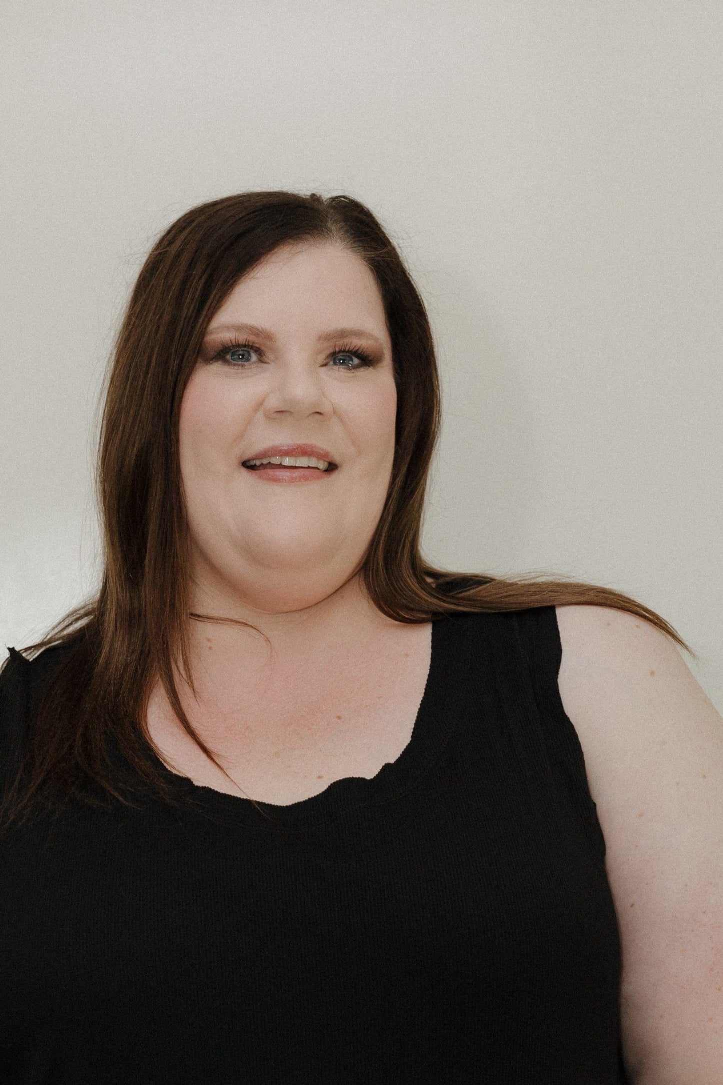 Woman with long brown hair wearing a black sleeveless top against a plain background