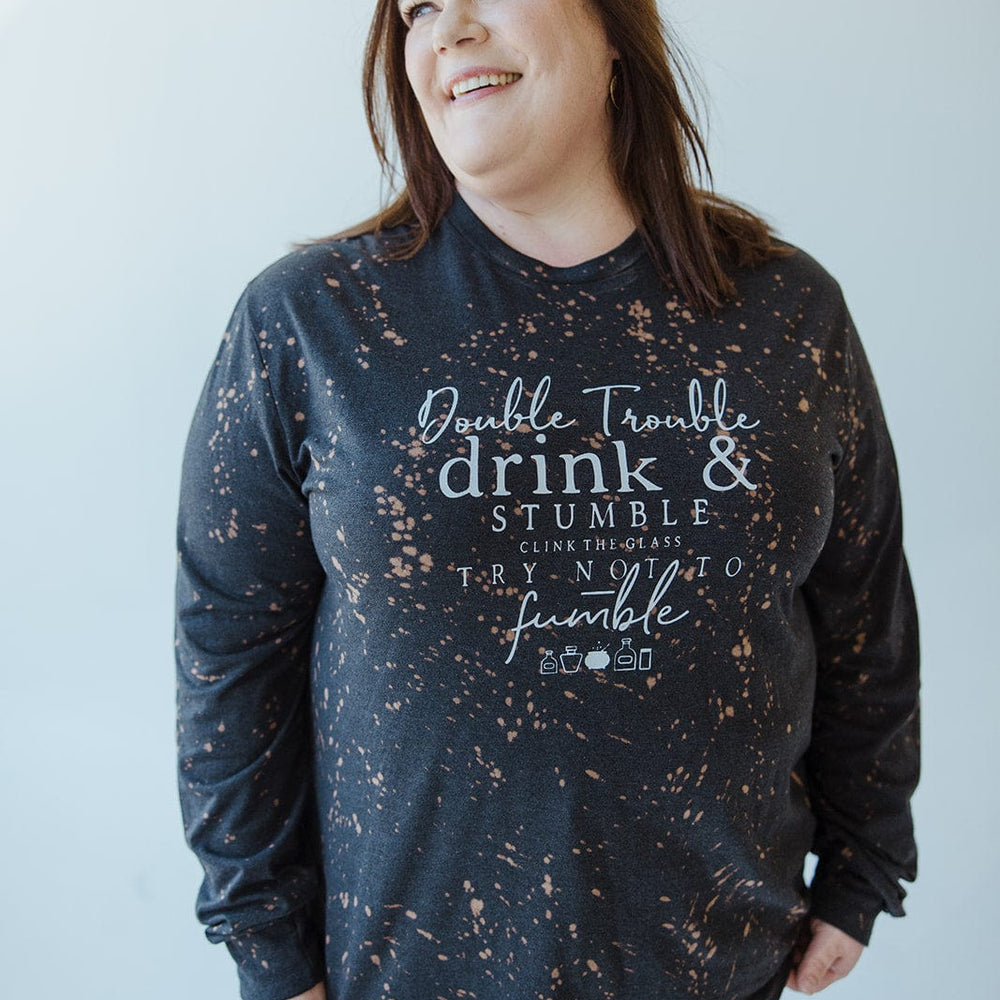 A person wearing the "DOUBLE TROUBLE DRINK AND STUMBLE CLINK THE GLASS TRY NOT TO STUMBLE" graphic tee from Love Marlow, featuring a black and orange speckled design with text and a unique splatter bleach pattern.