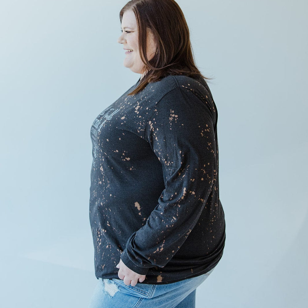 A person stands sideways, wearing the "DOUBLE TROUBLE DRINK AND STUMBLE CLINK THE GLASS TRY NOT TO STUMBLE" graphic tee by Love Marlow, paired with ripped jeans, set against a light gray background.